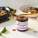 A jar of Terrapin Ridge Farms Thai Chili Guava Jam sits on a light surface with a gold spoon on top, accompanied by a lime wedge and plates of food in the background—ideal for adding sweet and spicy flavor to charcuterie boards.