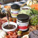 A charcuterie board with Terrapin Ridge Farms Raspberry Honey Mustard, cured meats, crackers, figs, artichoke, herbs, and decorative pumpkins arranged on a checkered tablecloth.