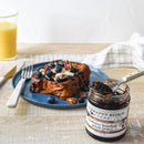 A 2oz jar of Terrapin Ridge Farms Blueberry Bourbon Pecan Jam with a spoon is in the foreground. Behind it, French toast topped with blueberries and pecans sits on a blue plate next to a glass of orange juice.