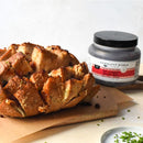 A sliced pull-apart bread loaf sits on a wooden board next to a jar of Terrapin Ridge Farms Hot Pepper Bacon Jam Quart, making an irresistible gourmet appetizer. Chopped chives are scattered nearby on the light surface.