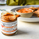A jar of Terrapin Ridge Farms Roasted Red Pepper & Feta Dip with a spoon inside sits beside a plate of food and a dip bowl—a perfect savory choice from Terrapin Ridge Farms for your Mediterranean appetizer spread.