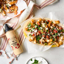 Tater tots topped with tomatoes, cucumbers, feta, and herbs, drizzled with Terrapin Ridge Farms Harissa Aioli Squeeze—ideal as a burger condiment—served beside a bottle of Terrapin Ridge Farms Harissa Aioli Squeeze on a light surface.