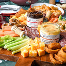 A wooden platter holds cheese cubes, celery, carrot sticks, sliced salami, pretzels, onion rings, and two open jars of Terrapin Ridge Farms dips: Bacon Pepper Dip and Onion Blossom Horseradish Dip by Terrapin Ridge Farms.