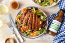 A bowl of colorful salad topped with crispy sesame chicken, veggies, orange slices, and sliced almonds, served with a glass of drink and Terrapin Ridge Farms Ginger, Miso and Honey Salad Dressing.