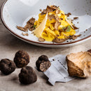 A plate of tagliatelle with truffle shavings and cheese sits on a beige tablecloth, surrounded by whole truffles, a slicer, and SALTOPIA Infused Sea Salts' Saltopia Parmesan Black Truffle Seasoning: GODFATHER in the foreground.