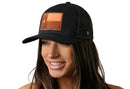 A woman with long brown hair smiles in a Haka Hat Texas Flag Trucker Hat Leather, a black snapback by Haka Hat featuring a rectangular brown leather patch with a star on the front, set against a plain white background.