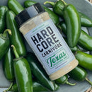 An 11 oz jar of Hardcore Carnivore Texas Jalapeño Salt Seasoning (Limited Release) from Hardcore Carnivore, a bold barbecue blend crafted with lively jalapeño flavor, rests on a gray plate surrounded by fresh green jalapeño peppers.