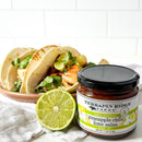 A plate of tacos with fresh lime and cilantro sits next to an open jar of Terrapin Ridge Farms Pineapple Chile Lime Salsa. A halved lime is in front, all set against a white background.