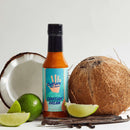 A bottle of Culture Hot Sauce Tahitian Dream from Culture Hot Sauce is showcased on a white surface, surrounded by a halved coconut, a whole coconut, lime wedges, and vanilla beans—beautifully expressing the tropical character of this hot sauce made with coconut, lime, and fragrant Tahitian vanilla.
