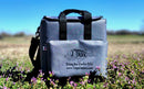 A Taiga Grey Back Pack Cooler by Taiga Coolers sits on grass and purple flowers under a blue sky, featuring black handles and a fish logo with “Living the Cooler Life” and TaigaCoolers.com.
