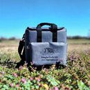 The Taiga Grey Back Pack Cooler by Taiga Coolers rests among purple wildflowers under a blue sky, its front pocket showing the brand’s logo and website, with sturdy black handles for easy carrying.