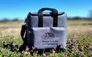 A Taiga Grey Back Pack Cooler by Taiga Coolers with black handles rests on grass among purple wildflowers, a blurred field and blue sky behind. “Living the Cooler Life” and a fish logo are on its front pocket.