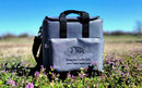 A Taiga Grey Back Pack Cooler by Taiga Coolers with black handles sits on grass and wildflowers under a blue sky, featuring a fish logo and “Living the Cooler Life www.TaigaCoolers.com”.