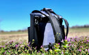 The Taiga Grey Back Pack Cooler by Taiga Coolers rests among purple wildflowers, set against a blurred grassy field and a clear blue sky.