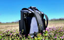 A Taiga Coolers Taiga Grey Back Pack Cooler rests on purple wildflowers under a clear blue sky, with blurred trees in the distance.