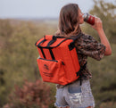 A person in a camouflage shirt and ripped denim shorts drinks from a bottle while carrying a Taiga Orange Back Pack Cooler by Taiga Coolers outdoors, surrounded by trees.