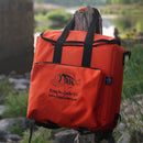 A Taiga Orange Back Pack Cooler with black straps hangs on a wooden post outdoors by water and greenery, showing the Taiga Coolers logo, website, and the slogan Living the Cooler Life.