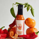 A bottle of Culture Hot Sauce Tangerine Dream by Culture Hot Sauce is displayed on a light surface with a white background, surrounded by tangerines, red chili peppers, green leaves, and peppercorns—beautifully capturing the vibrant spirit of this citrus hot sauce.