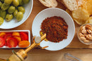A wooden board featuring green olives, sliced bread, potato chips, cherry tomatoes, and almonds surrounds a white bowl filled with World Spice A Taste of Portugal—a signature paprika and garlic spice blend from World Spice—capturing the rich flavors of Portuguese family traditions. An oil dispenser is also partially visible.