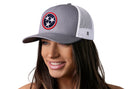 A smiling woman wears the Haka Hat Tennessee Flag Trucker Hat, a gray and white snapback featuring a circular patch with three white stars on a blue background surrounded by a red border.