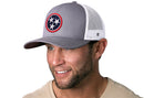 A man with a trimmed beard and beige shirt smiles while wearing the Haka Hat Tennessee Flag Trucker Hat in gray and white, featuring a circular patch with three white stars on a blue background.
