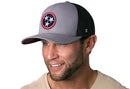 Smiling man with light stubble wears a Haka Hat Tennessee Flag Trucker Hat (Gray-Black TN Snapback) featuring a circular patch with three white stars on blue and red, plus a beige shirt, against a white background.