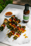 Colorful vegetable skewers with mushrooms, peppers, zucchini, and tomatoes are arranged on a marble surface next to a bottle of Terrapin Ridge Farms Ginger, Miso and Honey Salad Dressing and a small dish of dipping sauce.