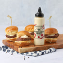 A bottle of Terrapin Ridge Farms Horseradish Sauce Squeeze sits on a wooden board alongside four mini roast beef sliders with skewers, scattered peppercorns, and a blue-patterned napkin.