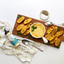 A wooden board featuring Terrapin Ridge Farms Avo Zingy Garlic Aioli in a white dish, cheesy dip, toasted bread slices, parsley garnish, pepper dish, silver spoon, kitchen towel, and a bottle of seasoning sauce.