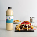 A quart of Terrapin Ridge Farms Avo Zingy Garlic Aioli sits beside a meat, slaw, and sauce sandwich with crinkle-cut fries. Salt and pepper shakers are visible in the background on a light-colored table.