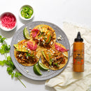 Three tacos topped with avocado, pickled onions, cheese, and herbs are served with lime wedges and Terrapin Ridge Farms Spicy Chipotle Garnishing Squeeze. Bowls of pickled onions, jalapeños, cilantro, and chipotle sauce complete the spread.