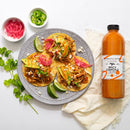 A plate with three tacos topped with shredded meat, pickled onions, cheese, and avocado is garnished with lime wedges. Next to it is a Terrapin Ridge Farms Spicy Chipotle Garnishing Aioli Quart, cilantro, and bowls of pickled onions and jalapeños.