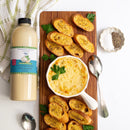 A quart-sized bottle of Terrapin Ridge Farms Avo Zingy Garlic Aioli sits beside a cheesy dip with parsley, toasted bread on a wooden board, a dish of salt and pepper, two spoons, and a white towel.
