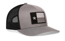 The Haka Hat Texas Flag Trucker Hat is a gray-black snapback with a mesh back and front patch featuring a stylized black, gray, and white Texas flag with one white star—an understated take on the classic design.