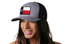 A smiling woman wears the Haka Hat Texas Flag Trucker Hat in gray and black, a snapback by Haka Hat featuring a Texas state flag patch on the front. The background is plain white.