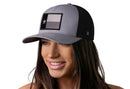 A woman with long brown hair smiles against a white background, wearing the Haka Hat Texas Flag Trucker Hat in gray and black, featuring a grayscale flag patch.