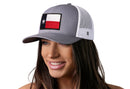 A woman with long brown hair smiles and looks to the side, wearing the Haka Hat Texas Flag Trucker Hat in gray and white, featuring a patch of the Texas flag. The stylish snapback is from Haka Hat.