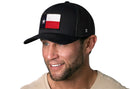 A smiling man in a beige shirt wears the Haka Hat Texas Flag Trucker Hat | Black TX Snapback by Haka Hat, featuring a flag patch on the front. He looks slightly to the side against a white background.