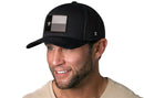 A man with a trimmed beard and short hair is smiling to the side, wearing a beige shirt and a Haka Hat Texas Flag Trucker Hat | Black TX Snapback featuring a black, white, and gray Texas flag patch on the front.