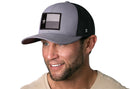A smiling man with a short beard wears a Haka Hat Texas Flag Trucker Hat in gray-black, featuring a patch of the black, gray, and white Texas flag with a star. He pairs it with a light beige shirt.