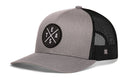 The Haka Hat Te(X)As Trucker Hat in gray-black features a circular patch with T, E, X, and A in a cross shape, classic snapback style, black mesh back, and is part of the HAKA signature trucker collection.