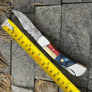 The Texan Knives Damascus Steel Pocket Knife with a Texas flag handle and 3" blade is displayed open on gray stone tiles next to a yellow measuring tape, showing it measures about 8 inches long. Includes leather sheath.