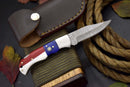 The Texan Knives Handcrafted Damascus Steel Pocket Knife features a Texas flag handle and comes with a brown leather sheath. Styled for collectors or EDC, it's displayed on a bundle with rope and autumn leaves, highlighting its rustic charm.