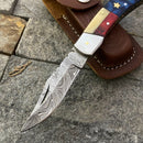 The Texan Knives Damascus Steel Pocket Knife with a Texas flag handle, featuring a 3" patterned blade, rests on stone beside its brown leather sheath.
