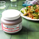 A quart jar of Terrapin Ridge Farms Thai Chili Guava Jam rests on a green table beside a plate of herb-garnished vegetables with lime wedges—this sweet-and-spicy jam from Terrapin Ridge Farms is perfect for cheese boards. Glasses of water appear in the background.