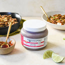 A quart jar of Terrapin Ridge Farms Thai Chili Guava Jam sits on a light surface, surrounded by prepared food, a gold spoon in a small bowl, and lime wedges—an ideal sweet-and-spicy cheese board topper.