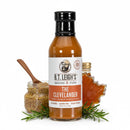A bottle of B.T. Leigh’s Mustard Maple Rosemary BBQ Sauce surrounded by a small jar of whole-grain mustard with a wooden spoon, a maple-leaf-shaped bottle of syrup, and sprigs of fresh rosemary.