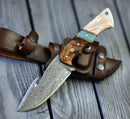 The Texan Knives Premium Damascus Guthook Skinning Knife features a patterned steel blade and a rosewood & bone handle, displayed on its brown leather sheath atop a wooden surface.