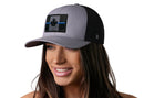 A woman with long brown hair smiles while wearing the Haka Hat Thin Blue Line Trucker Hat, a gray-black Canadian Maple Leaf tactical snapback by Haka Hat, which stands out against the plain white background.