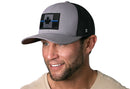A smiling man in a beige shirt wears the Haka Hat Thin Blue Line Trucker Hat by Haka Hat, a gray-black snapback featuring an embroidered Canadian maple leaf flag with a blue line, posed against a white background.